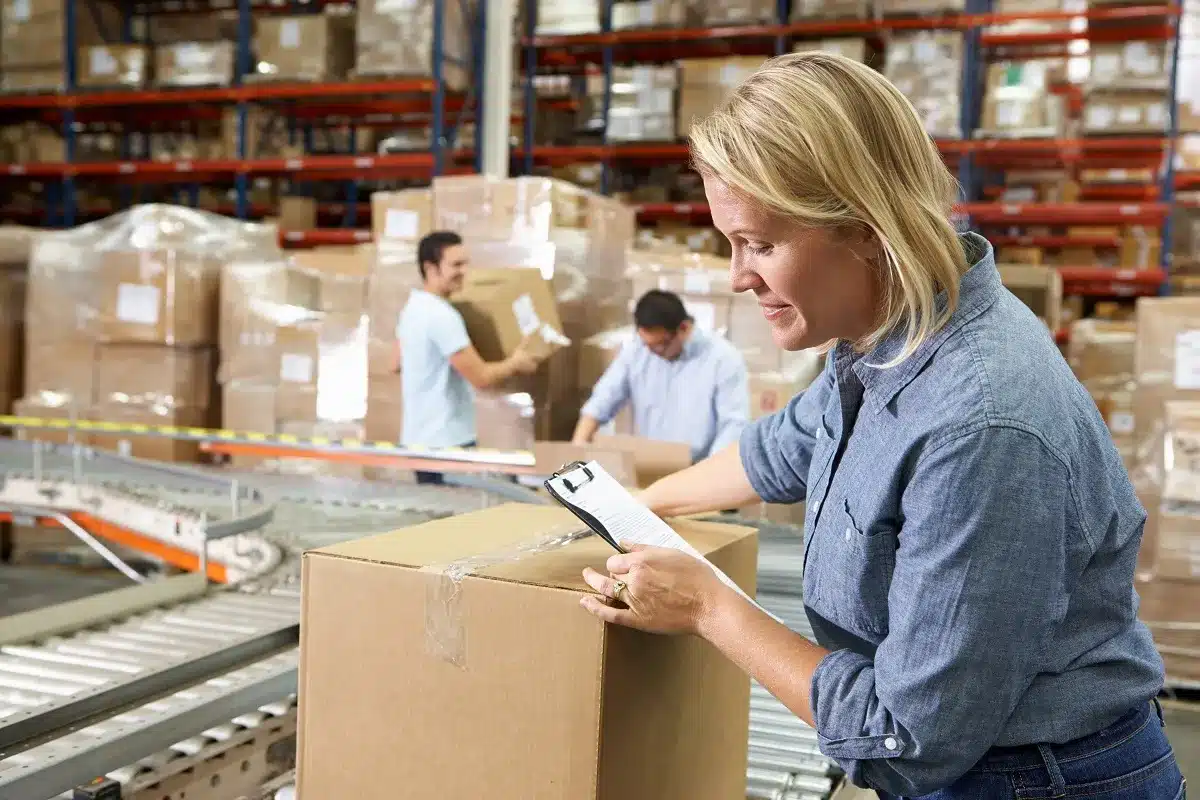 A woman checks a clipboard while inspecting a box on a conveyor belt in a warehouse. In the background, two men handle more boxes, utilizing cost reduction strategies. The scene suggests an active warehouse environment focused on operational efficiency.