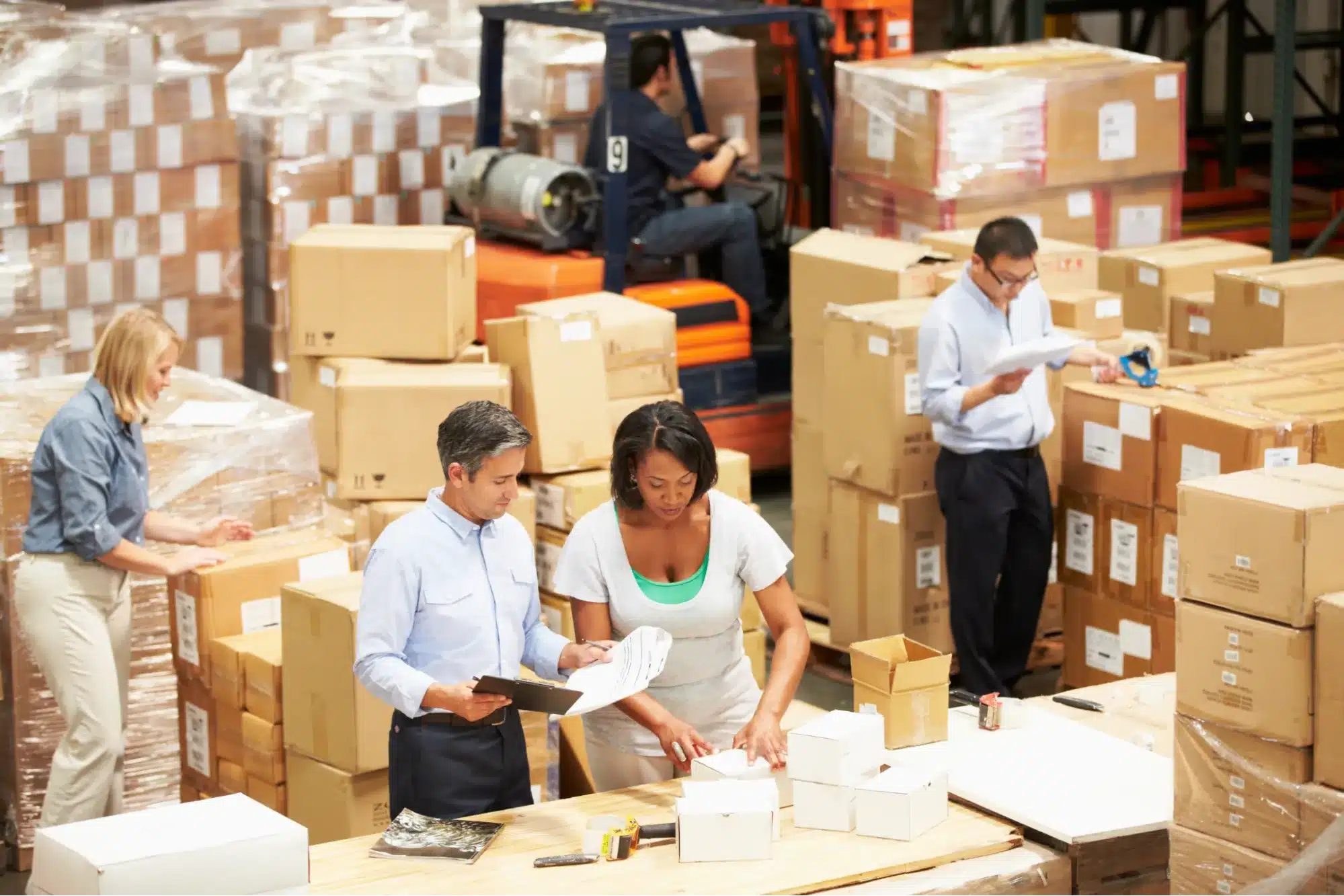 Four people are working in a warehouse, handling boxes and paperwork with a focus on enhancing efficiency. One person operates a forklift in the background, streamlining the workflow while the team integrates custom kitting services seamlessly into their routine.