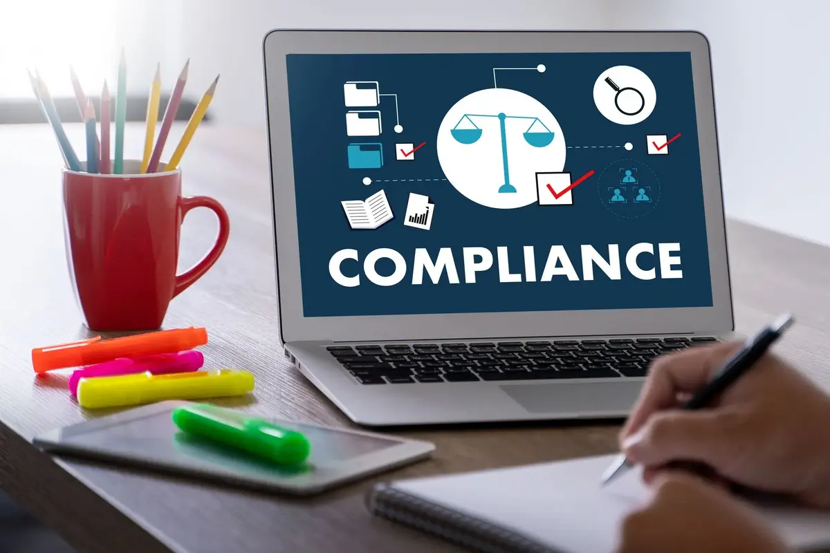 A person writes in a notebook at a desk with a laptop displaying a "COMPLIANCE" graphic, surrounded by colored markers, pencils, and a red mug—reflecting the organization's focus on HealthCare Logistics.
