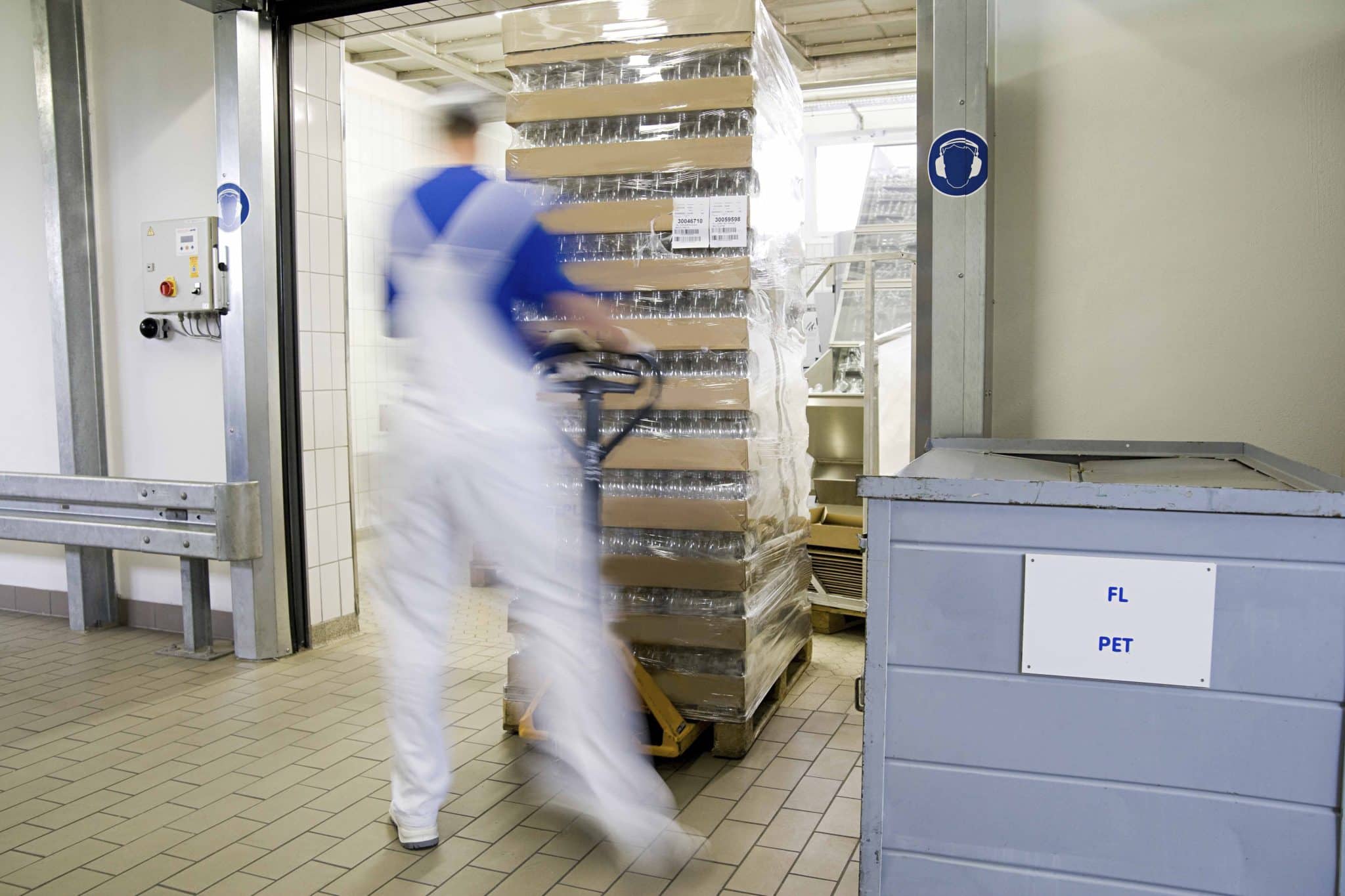 A worker in a white uniform navigates a manual pallet jack, moving a stack of boxes wrapped in plastic through a doorway in this tiled cold storage setting, efficiently managing seasonal demand.